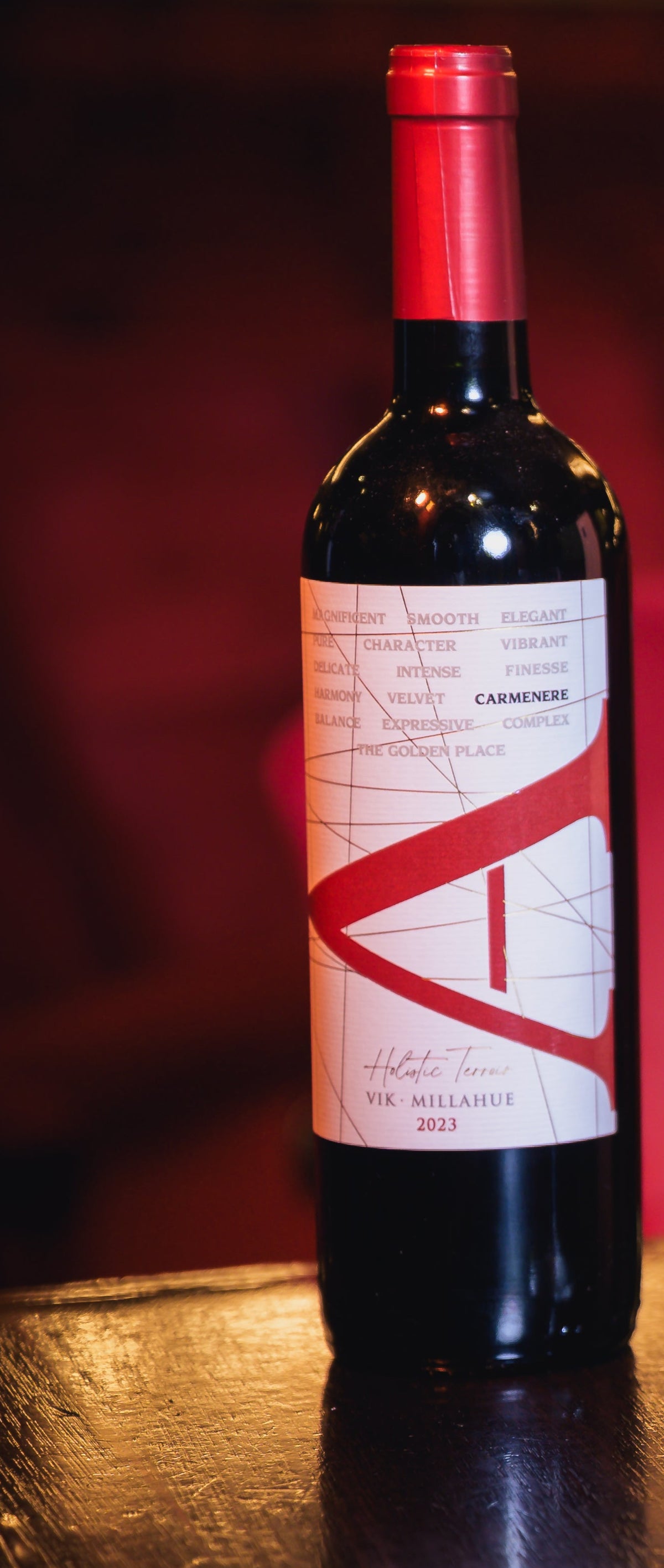 A bottle of red wine with a white and red label stands next to a clear wine glass on a dark wooden table, with a red upholstered venue background and a wooden box partially visible on the right.